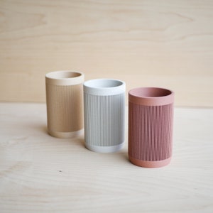 May include: Three cylindrical containers with a ribbed texture. The containers are beige, light gray, and light pink.