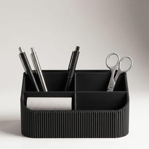 May include: A black desk organiser with a ribbed exterior texture featuring four distinct compartments. The organiser holds two black pens, one silver pen, and a pair of silver scissors, with a roll of white paper tape in the front left section.