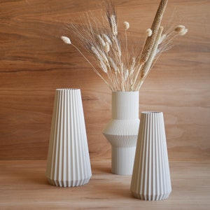 May include: Three white ceramic vases with a pleated design. The vases are arranged on a wooden surface. The tallest vase is filled with dried grasses and wheat.