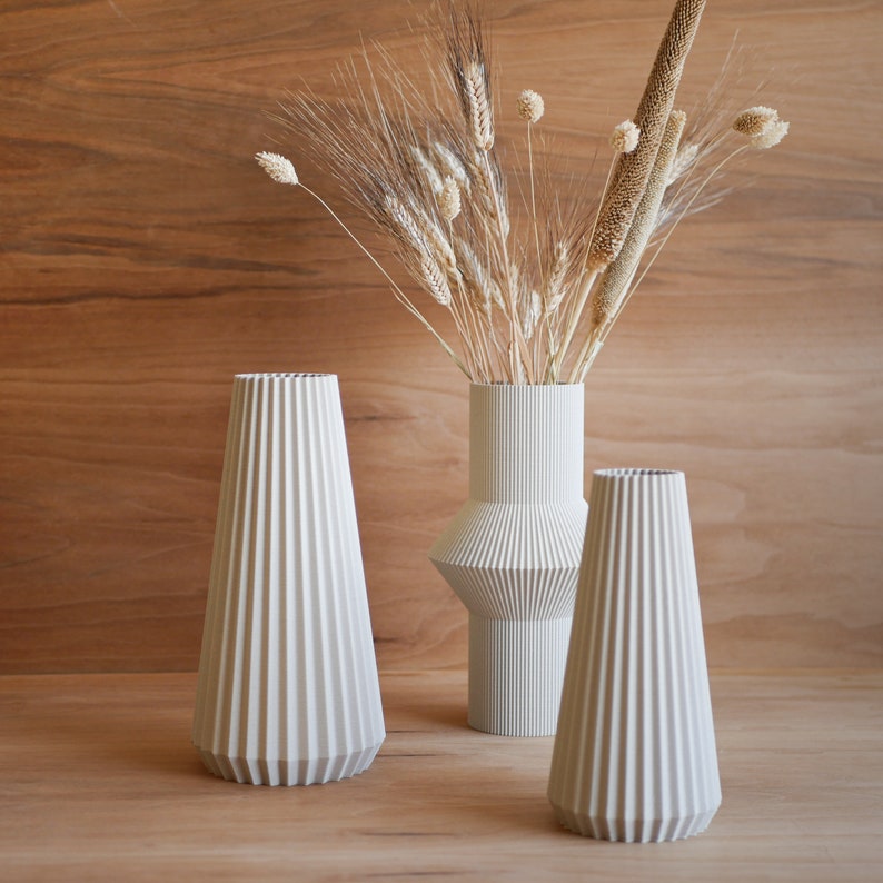 May include: Three white ceramic vases with a pleated design. The vases are arranged on a wooden surface. The tallest vase is filled with dried grasses and wheat.