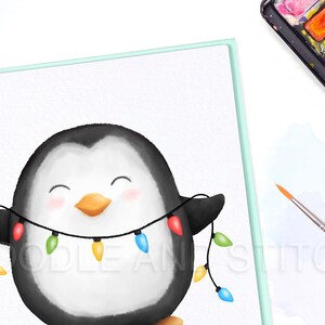 May include: A watercolor illustration of a penguin wearing a string of colorful Christmas lights. The penguin is smiling and has a happy expression. The illustration is drawn on a white page in a spiral bound notebook.