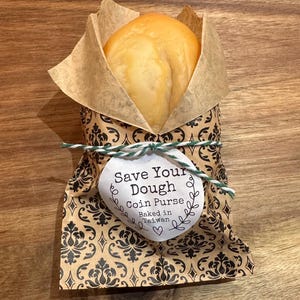 May include: A round, golden-brown coin purse shaped like a bread roll, wrapped in brown paper with a black damask pattern. A white circular tag is tied with green and white twine, reading: Save Your Dough Coin Purse Baked in Taiwan.