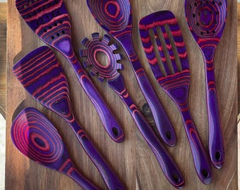 May include: A set of seven purple and red wooden kitchen utensils, including a slotted spoon, spatula, and serving spoon, arranged on a wooden cutting board. The utensils have a unique wood grain pattern.