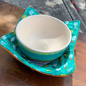 May include: A teal blue and orange bowl cozy with a pineapple pattern. The cozy is handmade and has the text "100% Cotton Handcrafted" on the bottom.