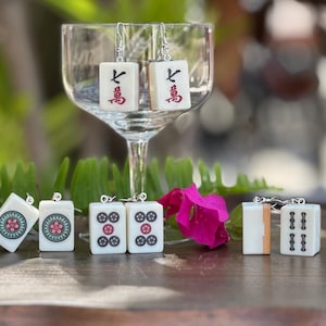May include: A collection of white Mahjong tile earrings with various symbols and characters in red and black. Some earrings are displayed in a wine glass, others are arranged on a wooden surface with greenery and a pink flower in the background.