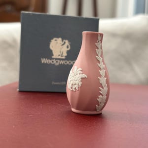 May include: A small, pink Wedgwood vase with white floral relief detailing. The vase has a narrow neck and a rounded body. A dark blue box with the Wedgwood logo is in the background.