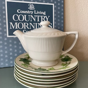 May include: A white ceramic teapot with a lid sits atop a stack of saucers with green ivy leaf designs. The saucers have a green border. A book titled "Country Living COUNTRY MORNINGS" is in the background.