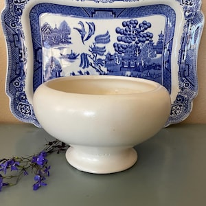May include: A white, footed bowl sits in front of a blue and white patterned tray. The tray has a scalloped edge and a landscape design. A sprig of purple flowers is in the foreground.