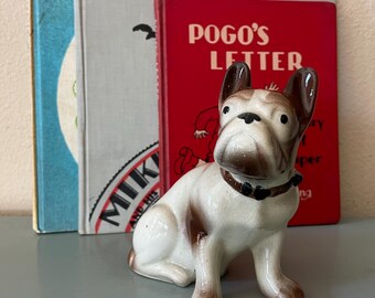 May include: A ceramic French bulldog figurine with a white body, brown patches on the ears and legs, and a brown collar. The dog is sitting in front of three books, including one with a red cover featuring the text POGO'S LETTER in white.