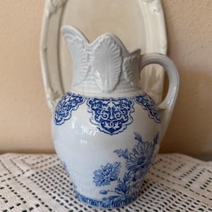 May include: A white ceramic pitcher featuring a blue floral pattern and intricate scrollwork around the upper rim. The pitcher has a curved handle and a molded leaf design at the top of the spout, with a glossy finish and a rounded body shape.