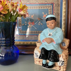 May include: A blue glass vase holds orange and yellow flowers. A ceramic figurine of a person in a light blue outfit and black pants sits on a woven basket. The figurine has a blue head covering and is holding their hands together.