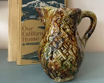 May include: A small, vintage ceramic pitcher with a woven basket pattern in shades of brown, green, and cream. The pitcher has a curved handle and a spout. Behind the pitcher are two old books, one with the title "Our California Home".