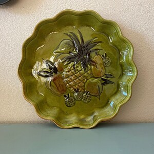 May include: A green ceramic serving platter with a scalloped edge. The center features a raised relief design of a pineapple, pears, and other fruits. The fruits are painted in shades of brown, green, and yellow.