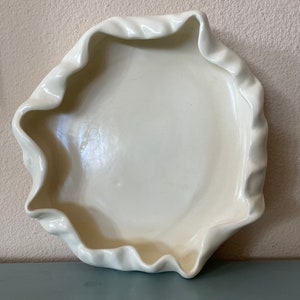 May include: Off-white ceramic bowl with a ruffled edge. The bowl has a shallow, rounded interior and a wavy, textured rim. The color is a creamy, pale hue, and the bowl appears to be handmade.