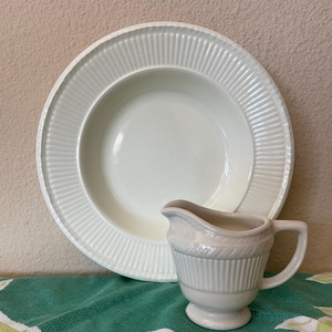 May include: A white ceramic serving bowl and creamer set. The bowl has a wide rim with vertical ridges, and the creamer has a handle and a small spout. The set is on a green and white floral patterned cloth.