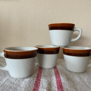 May include: Four white ceramic coffee mugs with a brown band around the top. Each mug has a handle and is stacked on a white and red striped cloth. The mugs are a simple, classic design.