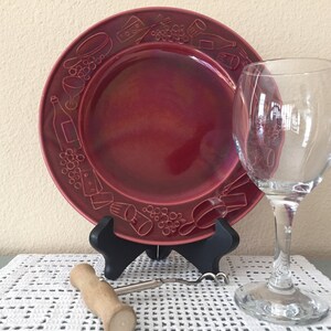 May include: A burgundy-colored plate with embossed wine-themed designs, a clear wine glass, and a corkscrew. The plate is displayed on a black stand, and the items are set on a white crocheted tablecloth. The plate's border features grapes, bottles, and cheese.