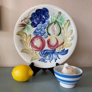 May include: A white ceramic plate with a hand-painted fruit design, including grapes, figs, and apples. A yellow lemon and a small bowl with blue stripes and garlic are also present. The plate is on a black stand.