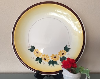 May include: A large, round decorative plate with a white center and a yellow and brown rim. The plate features a hand-painted floral design of yellow daisies and green leaves. A small white pitcher with red flowers sits in front of the plate.
