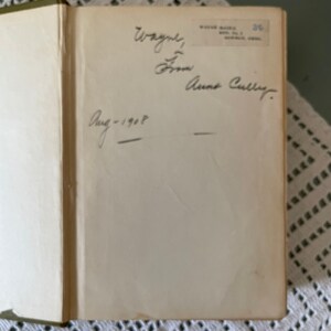 May include: Open book with handwritten inscription: "Wayne, From Anne Cully. Aug-1908." The book's pages are off-white, and the binding is green. The book is resting on a white lace tablecloth.