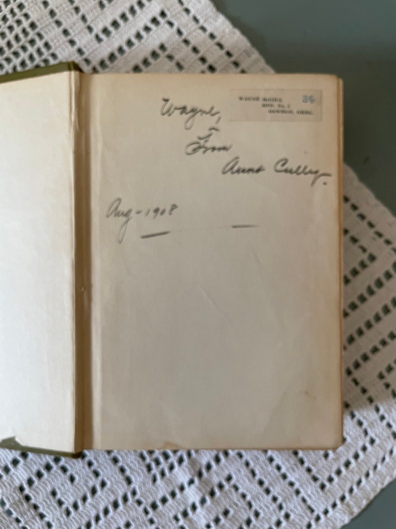 May include: Open book with handwritten inscription: "Wayne, From Anne Cully. Aug-1908." The book's pages are off-white, and the binding is green. The book is resting on a white lace tablecloth.