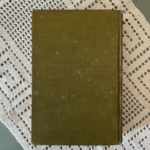 May include: An olive green book with a textured cover. The book is rectangular and appears to be vintage. The cover shows signs of wear and tear, with some discoloration. The book is resting on a white, crocheted doily.