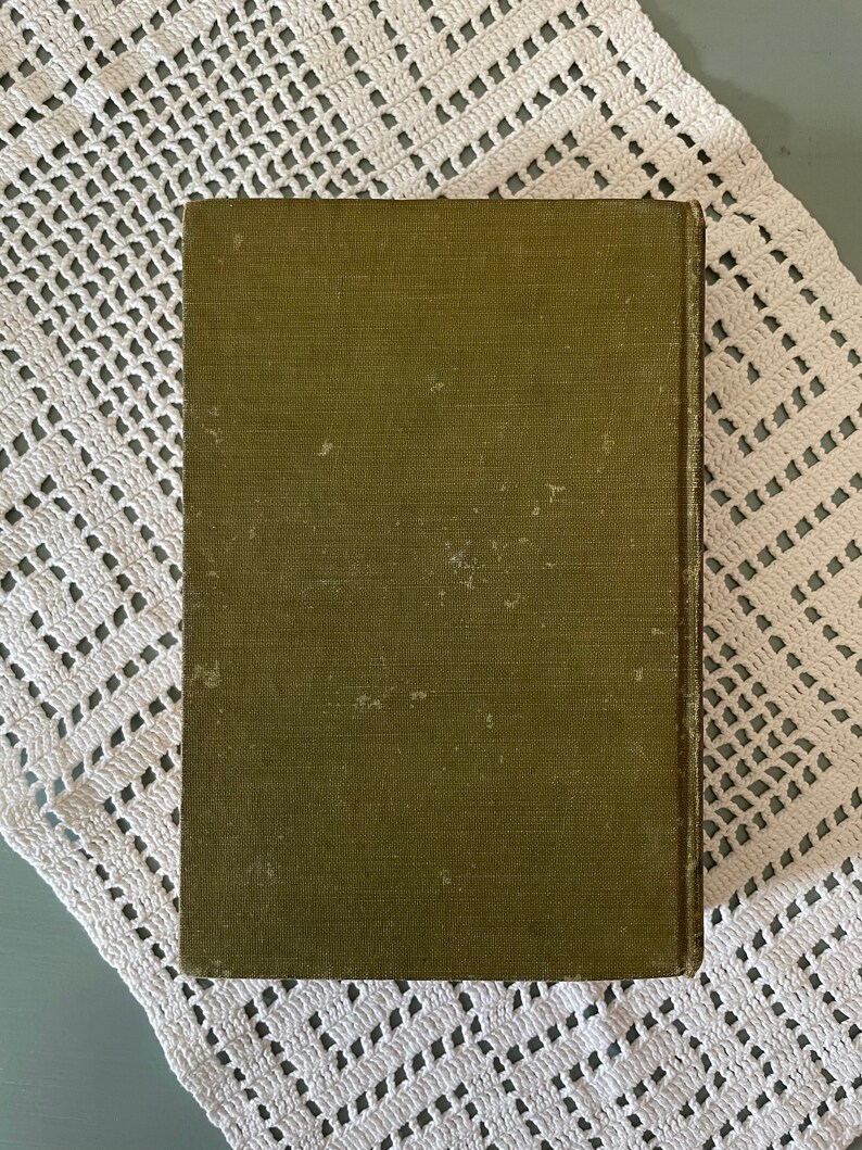 May include: An olive green book with a textured cover. The book is rectangular and appears to be vintage. The cover shows signs of wear and tear, with some discoloration. The book is resting on a white, crocheted doily.
