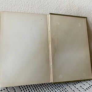 May include: An open book with aged, off-white pages. The spine is green, and the pages show signs of wear and age. The book is lying open on a white lace tablecloth.