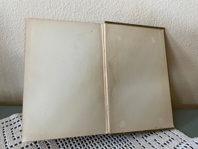 May include: An open book with aged, off-white pages. The spine is green, and the pages show signs of wear and age. The book is lying open on a white lace tablecloth.