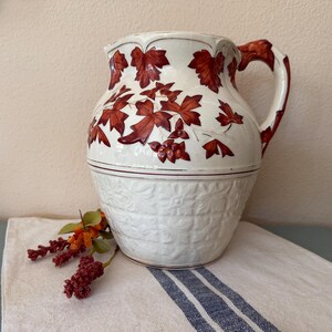 May include: A cream-colored ceramic pitcher featuring a raised, hand-painted pattern of dark red maple leaves on the upper section and a textured, embossed grid design on the lower half. The handle is also decorated with red leaf motifs.