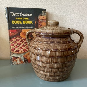 May include: A vintage brown ceramic cookie jar with a lid, featuring a textured brick pattern. The jar has two handles and sits next to a vintage cookbook titled "Betty Crocker's Picture Cook Book."