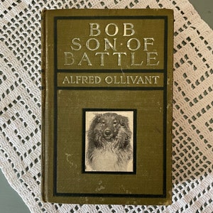 May include: An antique book titled "Bob, Son of Battle" by Alfred Ollivant. The cover is olive green with silver lettering and a black and white portrait of a dog. The book rests on a white, open-weave doily.