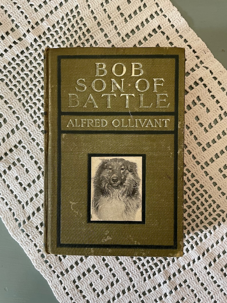 May include: An antique book titled "Bob, Son of Battle" by Alfred Ollivant. The cover is olive green with silver lettering and a black and white portrait of a dog. The book rests on a white, open-weave doily.