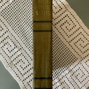 May include: A vintage book titled "Bob, Son of Battle" by Ollivant. The book's spine is olive green with black bands and embossed text. The book is resting on a white, open-weave, crocheted doily.
