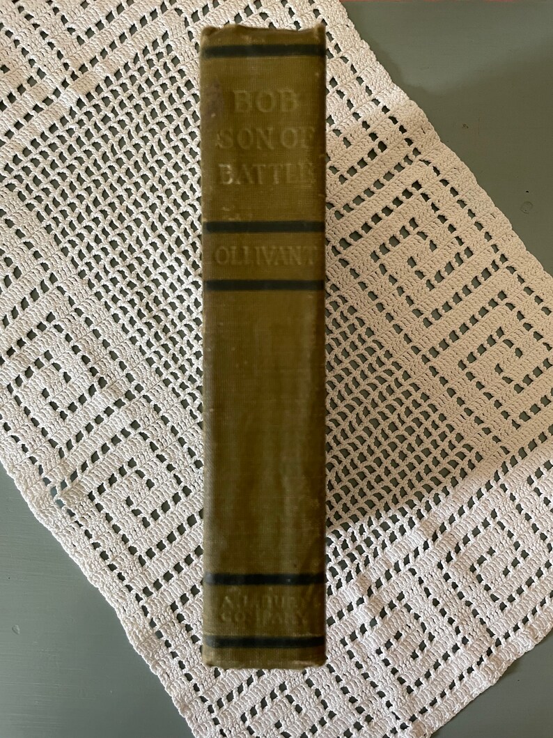May include: A vintage book titled "Bob, Son of Battle" by Ollivant. The book's spine is olive green with black bands and embossed text. The book is resting on a white, open-weave, crocheted doily.