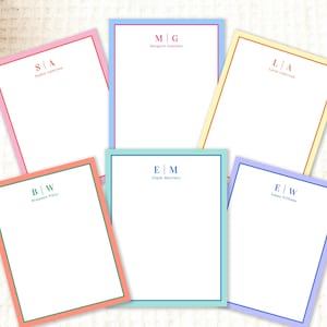 May include: A collection of personalized stationery with colorful borders. Each sheet features initials and a name in a complementary color to the border. The stationery is arranged on a light-colored surface, suggesting a stationery or gift shop.