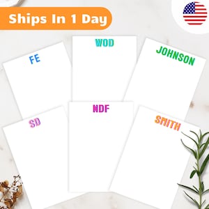 May include: White notepads with colorful text, including "WOD", "JOHNSON", "SMITH", "NDF", "SD", and "FE". The notepads are arranged on a marble surface with a sprig of greenery. An orange banner reads "Ships In 1 Day".