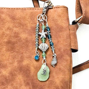 May include: A beach-themed purse charm with a lobster clasp. The charm features a sea glass pendant with the letter "B", a starfish, a seashell, and various blue and green beads. The charm is attached to a brown purse.