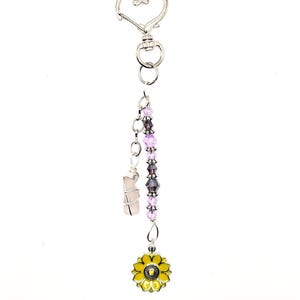 May include: A silver-toned keychain with a heart-shaped clasp. It features a chain with purple and clear beads, a wrapped pink crystal, and a yellow sunflower charm with black detailing. The keychain is designed for decorative use.