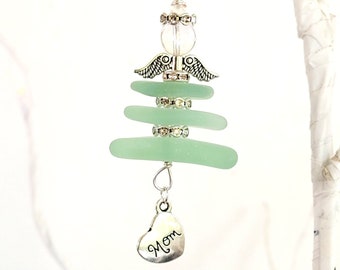 Pode incluir: Enfeite artesanal com um design de anjo de vidro do mar. O anjo tem uma cabeça de contas transparente, asas prateadas e um corpo de vidro do mar verde empilhado. Um pingente de coração prateado com a palavra "Mãe" pende na parte inferior. O enfeite é suspenso por um fio de prata.