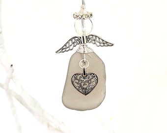 Pode incluir: Enfeite de anjo artesanal com corpo de vidro do mar cinza e um pingente de coração de filigrana prateada. O anjo tem asas prateadas, uma cabeça de contas transparentes e uma auréola de arame prateado. Um item decorativo.