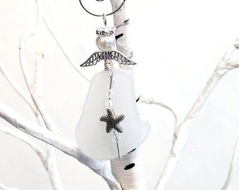 Pode incluir: Um enfeite de anjo artesanal feito de vidro do mar fosco. O anjo apresenta asas prateadas, uma cabeça de pérola e um pingente de estrela do mar. Ele pende de um fio decorativo prateado. O enfeite é exibido em um galho de árvore branco.