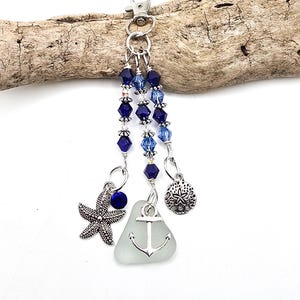 May include: A silver-toned metal keyring featuring a lobster clasp, three dangling strands of blue and light blue faceted crystal beads, a silver starfish charm, a silver sand dollar charm, and a piece of frosted sea glass with an anchor charm.