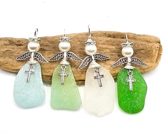 Pode incluir: Quatro enfeites de anjo de vidro do mar em tons de azul, verde e branco. Cada anjo tem uma cabeça de pérola, asas prateadas e um pingente de cruz. Eles são exibidos em um pedaço de madeira flutuante contra um fundo branco.