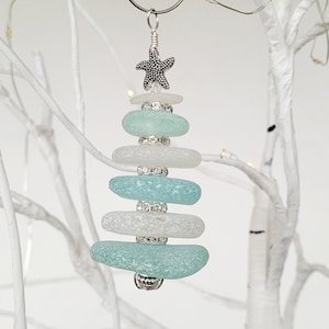 May include: A teal and white sea glass Christmas tree bauble with a silver starfish and silver beads. The bauble is hanging from a silver wire hanger.