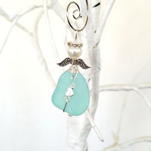 Puede incluir: Un adorno de ángel de vidrio azul claro con alas plateadas, una perla y un dije de corazón plateado. El adorno está colgado de un lazo de alambre plateado.