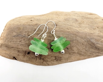 May include: Green sea glass earrings with silver hooks and small silver beads. Each earring has three stacked pieces of sea glass, creating a layered effect. The earrings are displayed on a piece of driftwood.