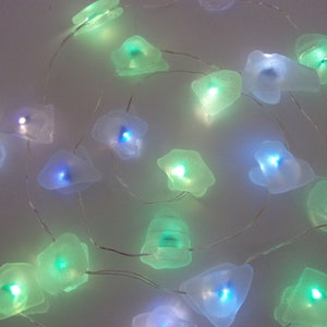 Puede incluir: Una cadena de luces decorativas con cubiertas translúcidas de forma irregular. Cada cubierta emite un brillo suave en tonos de azul y verde. Las luces están ensartadas en un alambre fino y plateado, creando un efecto caprichoso y etéreo.