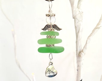 Pode incluir: Um enfeite de anjo artesanal com peças empilhadas de vidro do mar verde claro. O anjo tem asas prateadas, detalhes decorativos e um pingente de coração com a palavra "Mãe". O enfeite é pendurado por um fio de prata.