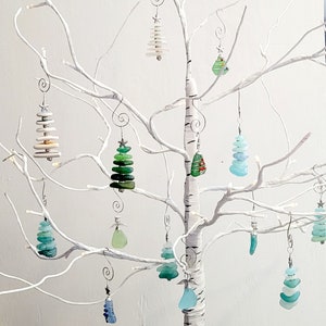 May include: A white twig tree with 12 glass ornaments shaped like Christmas trees. The ornaments are decorated with green, blue, and white glass pieces.